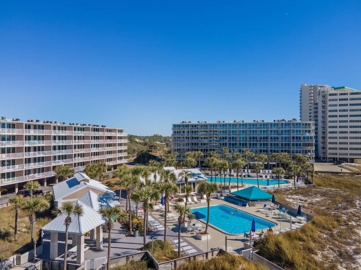 Dunes C605 – View of a vacation rental complex with swimming pools and palm trees under a clear blue sky.