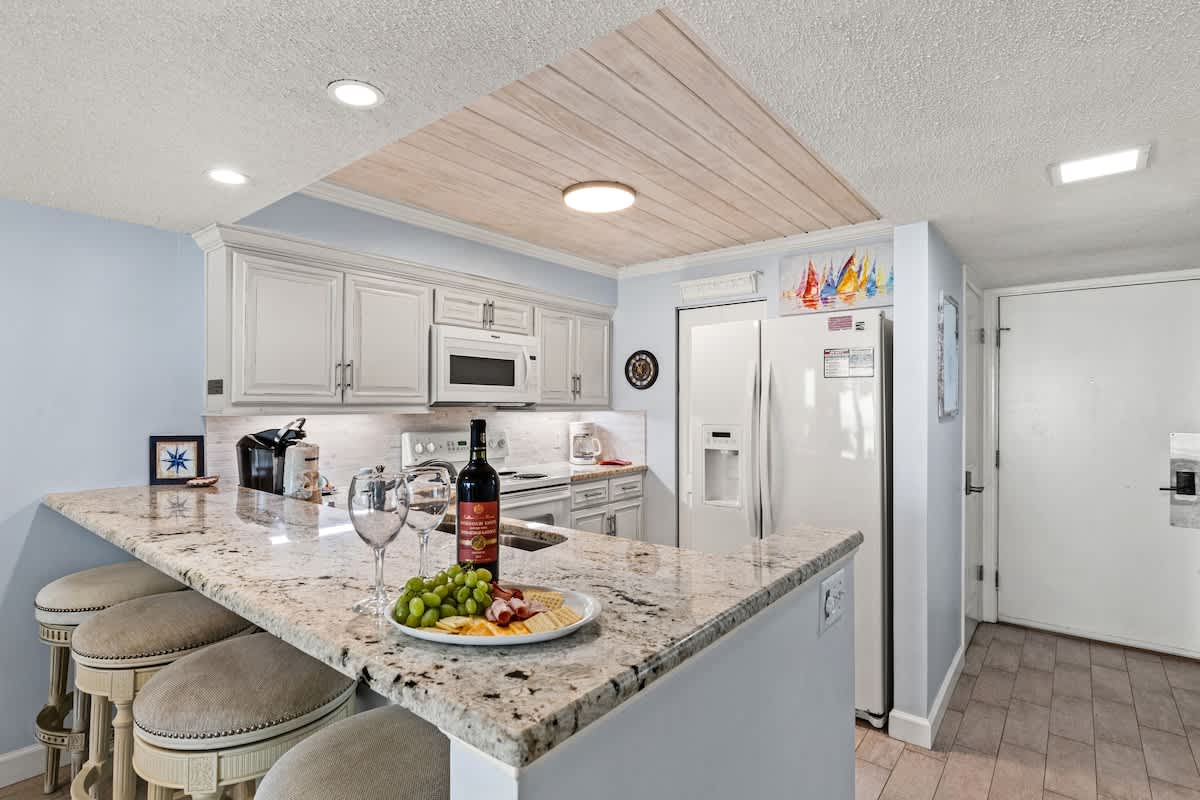 Dunes C605 – Modern kitchen with granite counter, bar stools, stainless steel appliances, and a decorative plate with snacks.