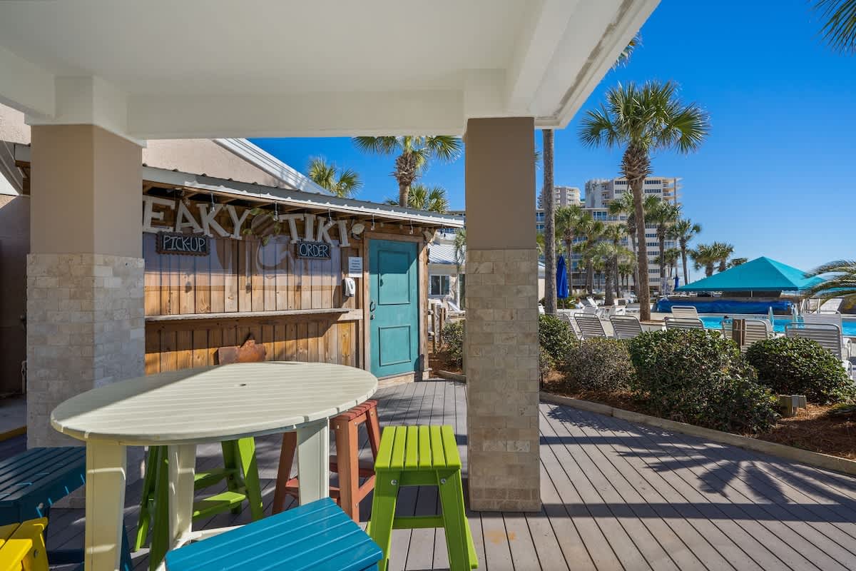 Dunes C605 – Outdoor area with colorful tables, Tiki bar, swimming pool, and palm trees in the background.