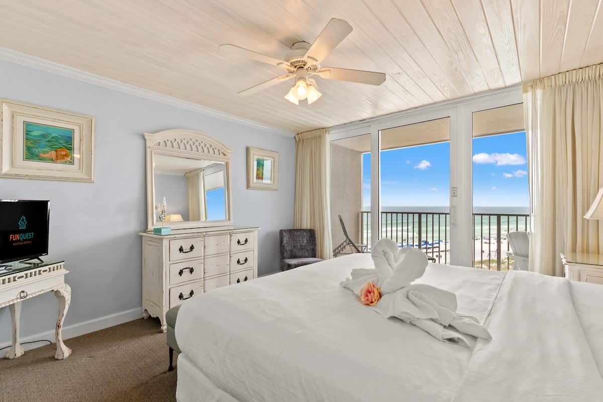 Dunes C605 – Bedroom with a white bed, dresser, TV, and ocean view through sliding glass doors.