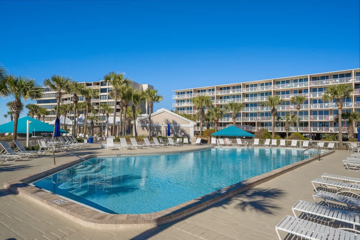Dunes C605 – Swimming pool surrounded by palm trees and lounge chairs, with multiple condominium buildings in the background.