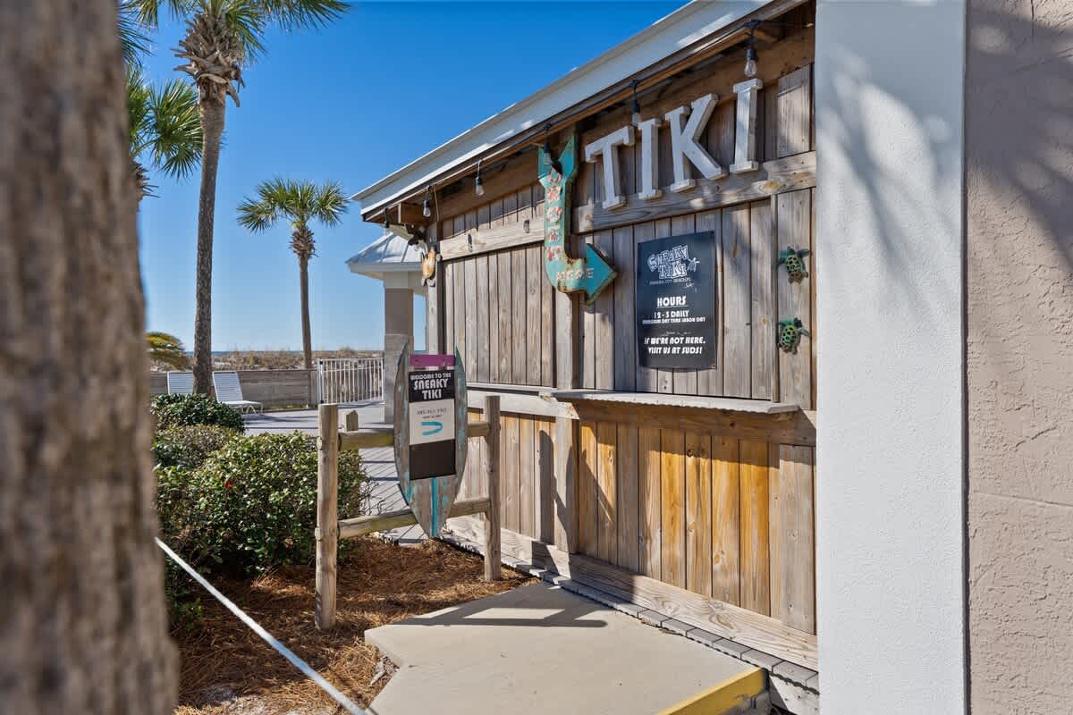 Dunes C605 – Tiki bar entrance with wooden signage, palm trees, and a view of the beach in the background.