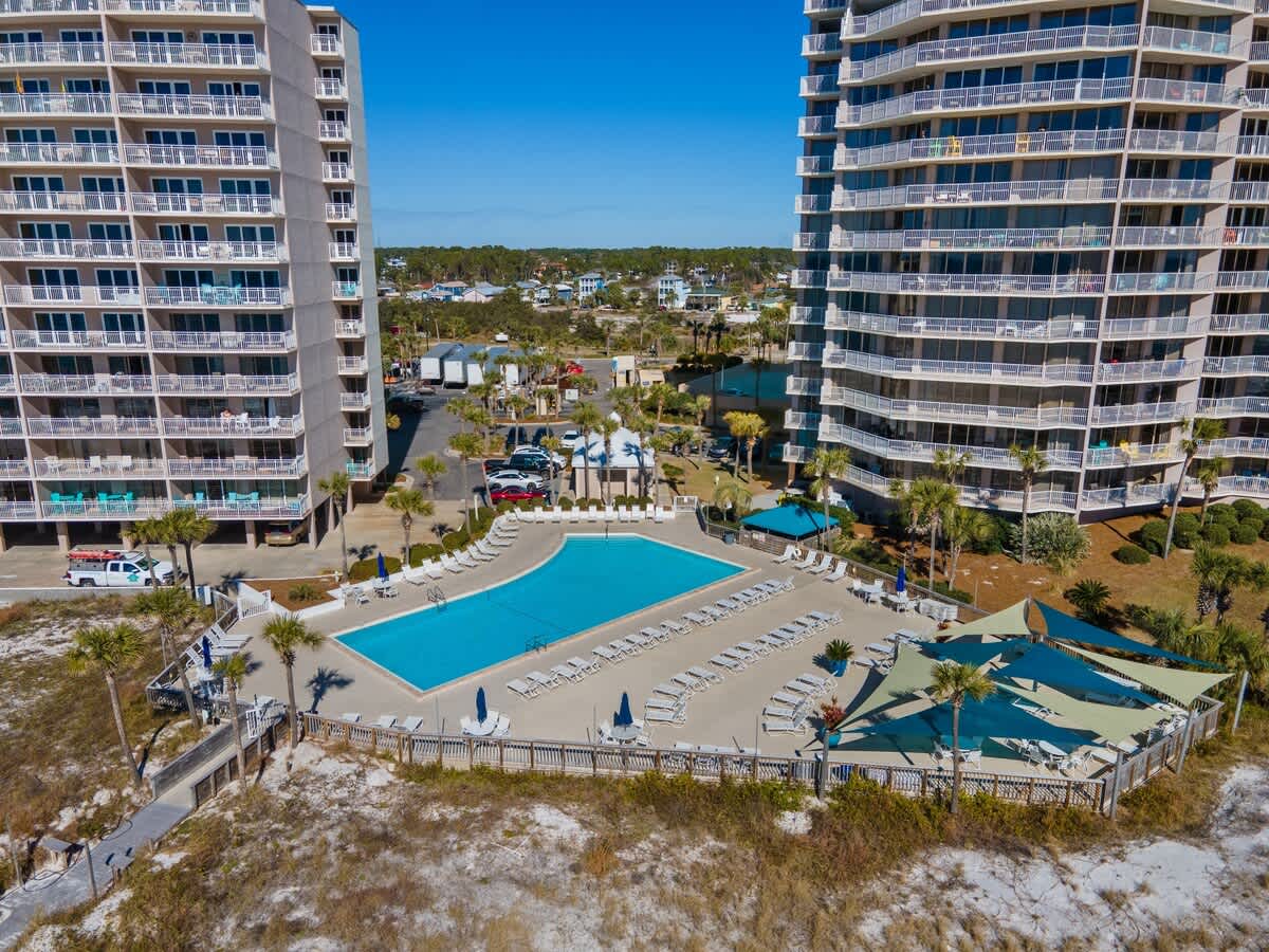 Dunes C605 – Aerial view of two tall buildings beside a swimming pool surrounded by lounge chairs and palm trees.