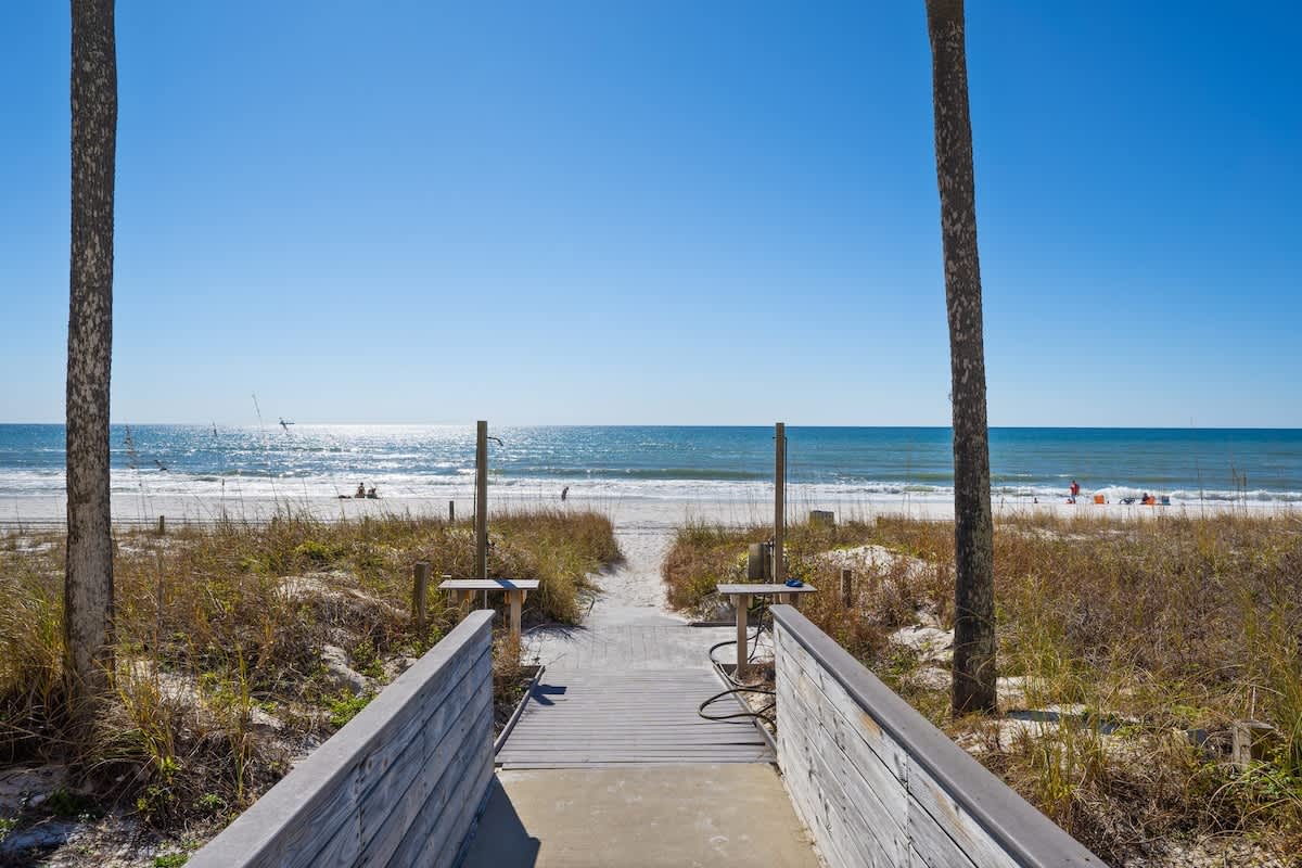 Dunes C605 – Pathway leading to the beach, bordered by grass and palm trees, with clear blue skies and ocean view.