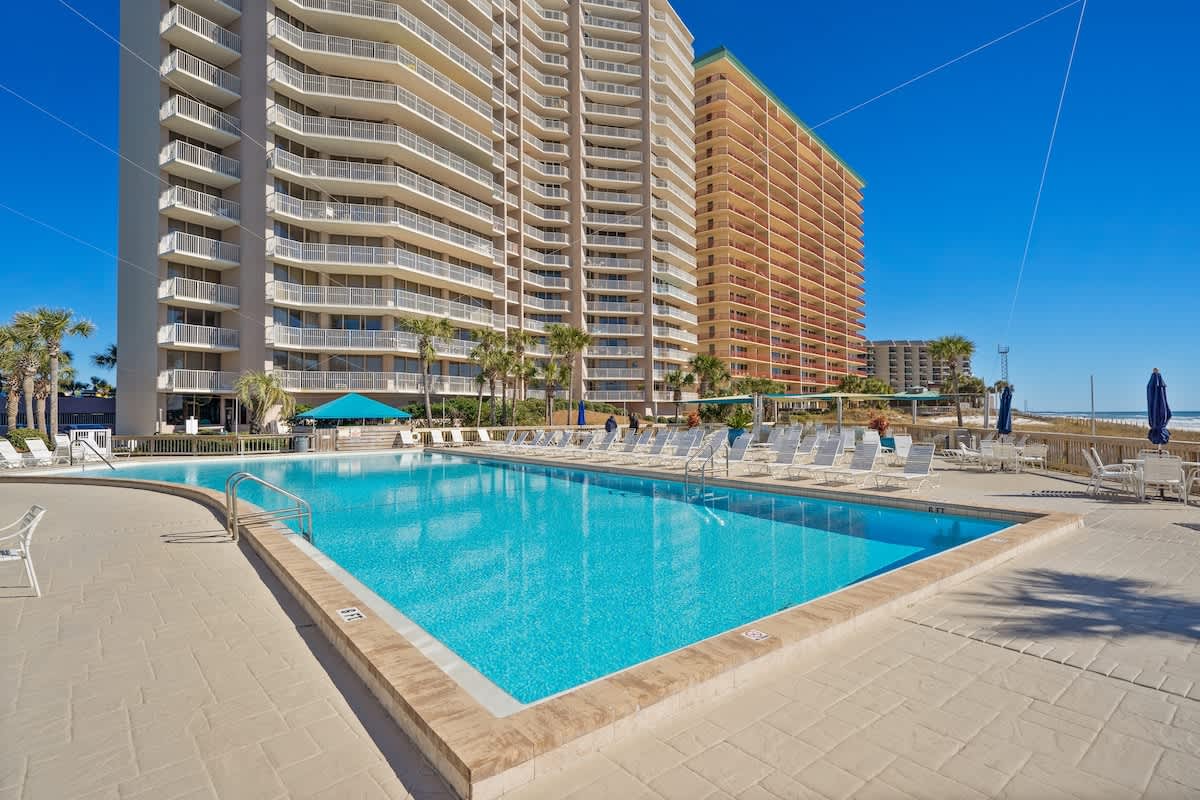 Dunes C605 – Swimming pool surrounded by lounge chairs, with tall buildings and palm trees in the background.