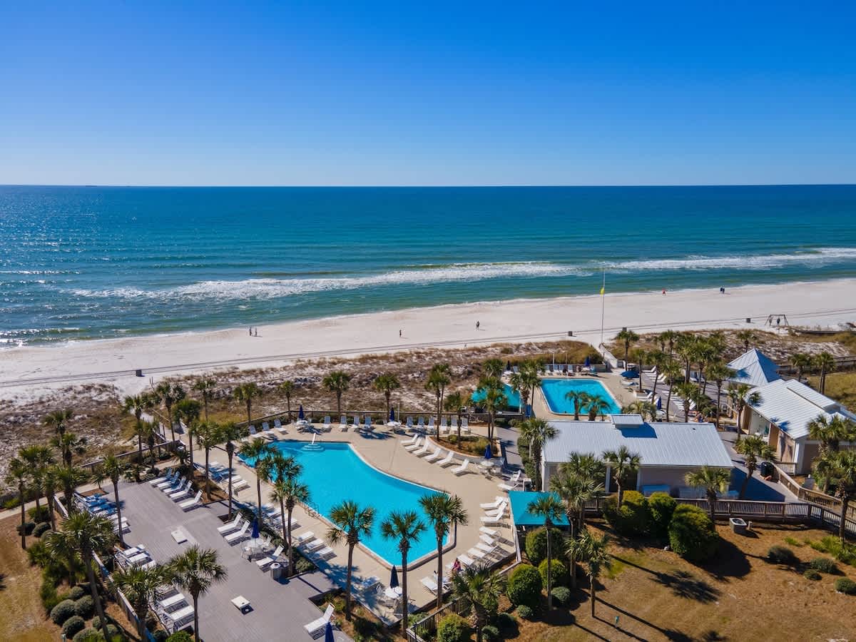 Dunes C605 – Aerial view of a beachside rental property featuring a swimming pool, palm trees, and a sandy beach.
