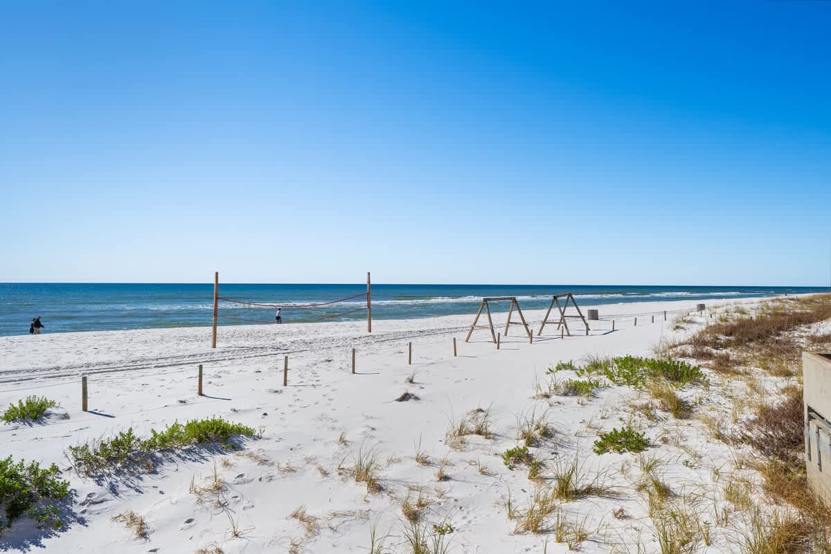 Dunes C605 – Beach view with volleyball net, swings, and sandy shoreline under a clear blue sky.