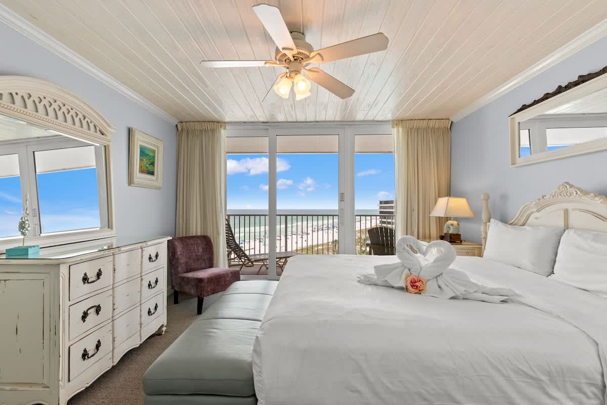 Dunes C605 – Bright bedroom with a king bed, seating area, dresser, and ocean view through large sliding glass doors.