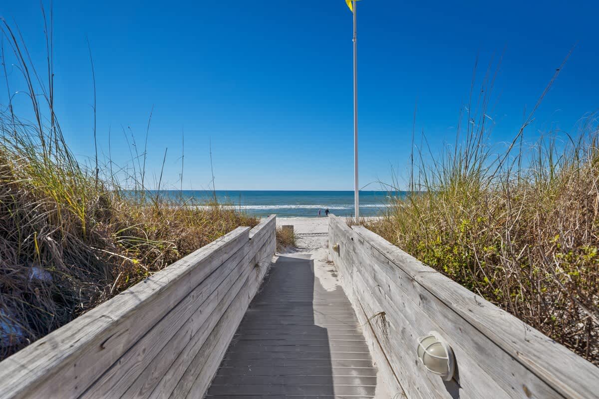 Dunes C605 – Pathway leading to the beach with grass on either side and a clear sky.