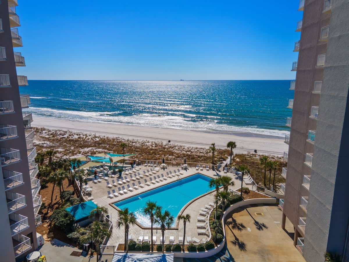 Dunes C605 – View of ocean, pool, and beach from a balcony with nearby palm trees and lounge chairs.