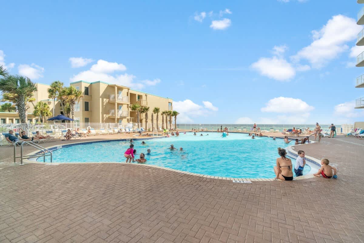 Tidewater 200 – Pool area with swimmers, lounge chairs, and palm trees, with a view of a building and blue sky.