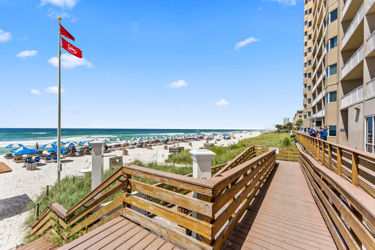 Tidewater 200 – Boardwalk leading to a beach with lounge chairs and umbrellas, ocean view, and nearby condominium building.
