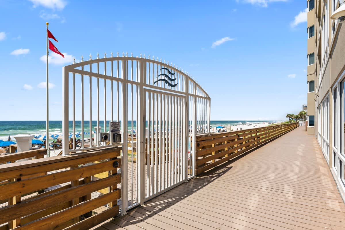 Tidewater 200 – Beachfront access gate leading to a wooden walkway and beach with umbrellas and ocean in view.