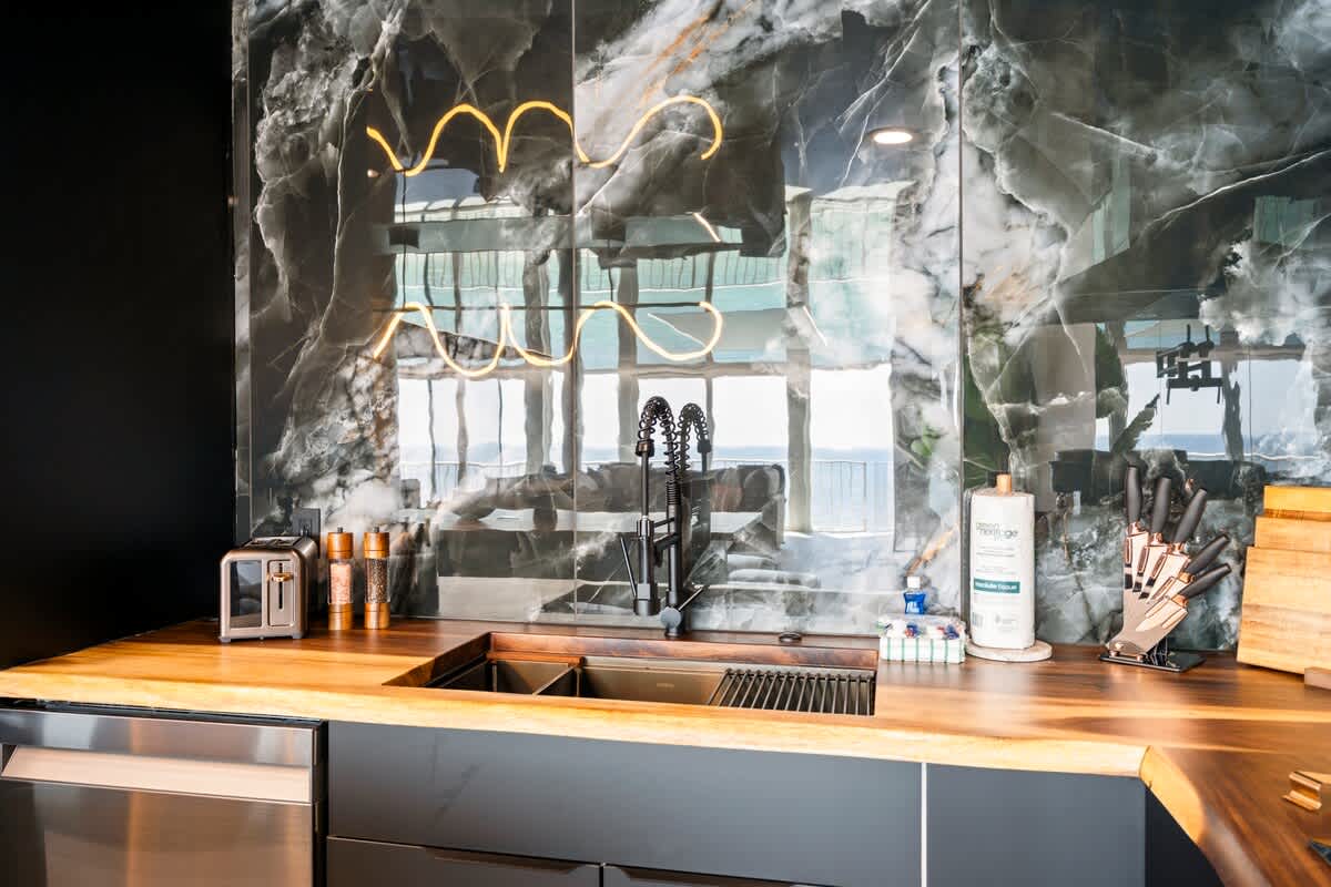 The Summit Penthouse 1501 – Modern kitchen with dark marble backsplash, wooden countertop, sink, and knife block, featuring a view beyond.