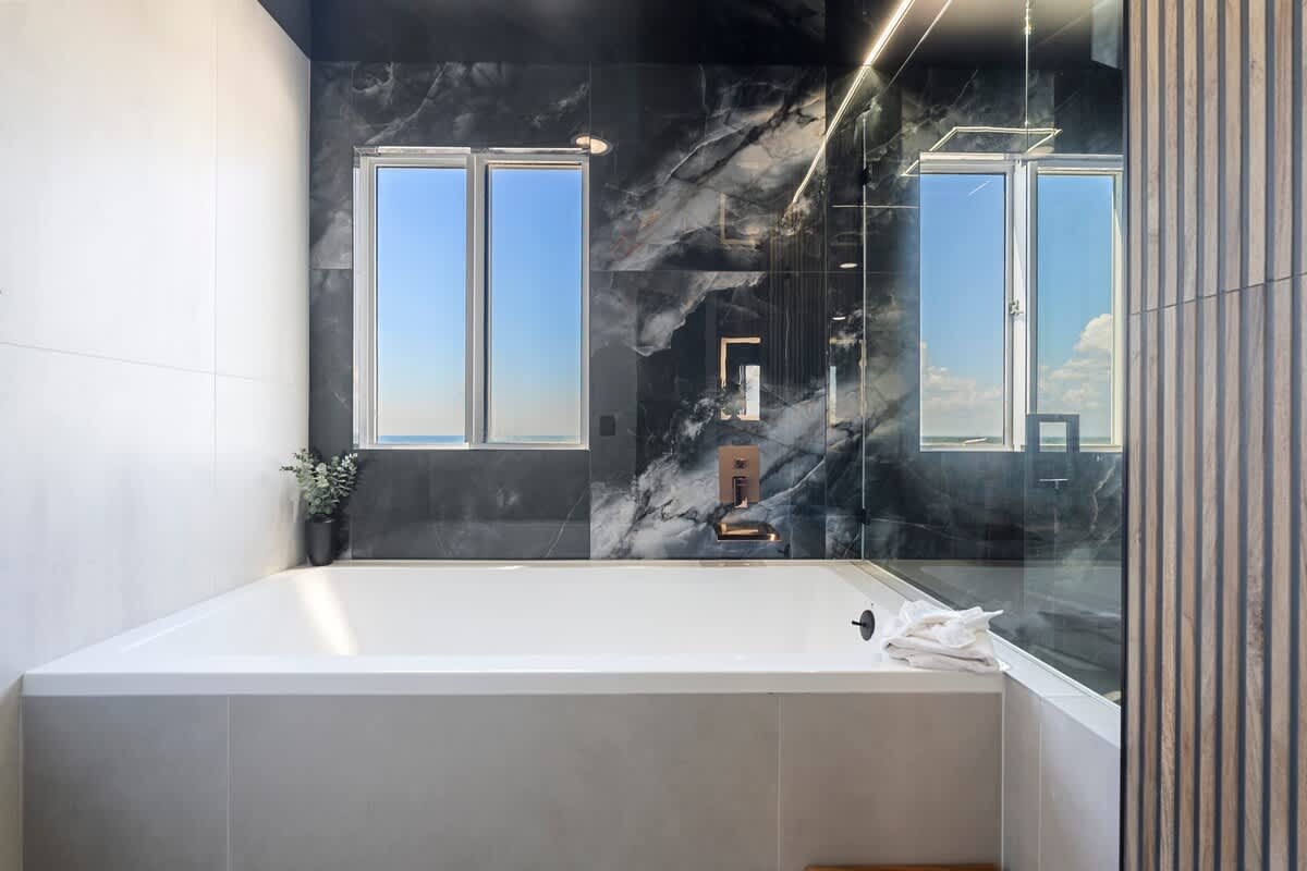 The Summit Penthouse 1501 – Modern bathroom with a large white tub, marble walls, and windows showcasing a scenic view.