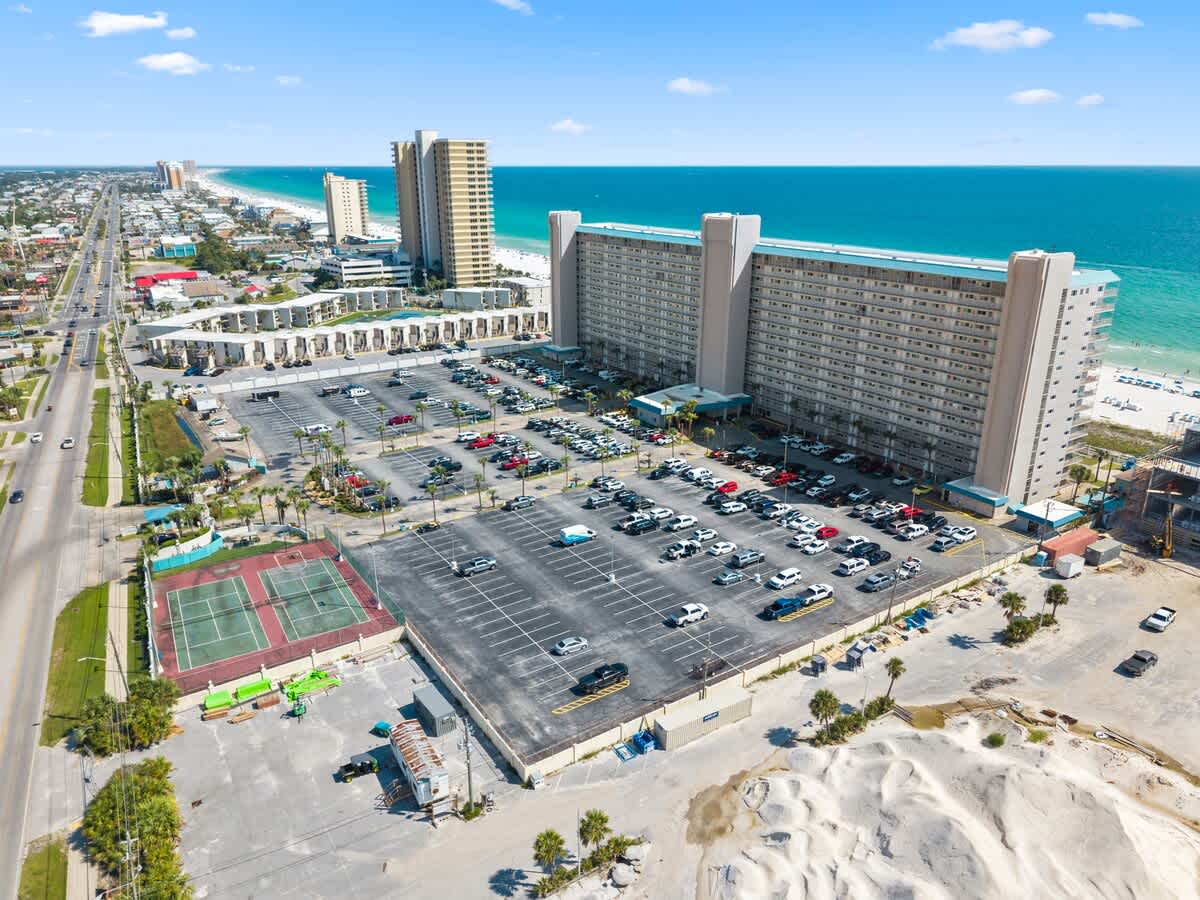 The Summit Penthouse 1501 – Aerial view of a large beachfront hotel, parking lot, tennis courts, and coastline.