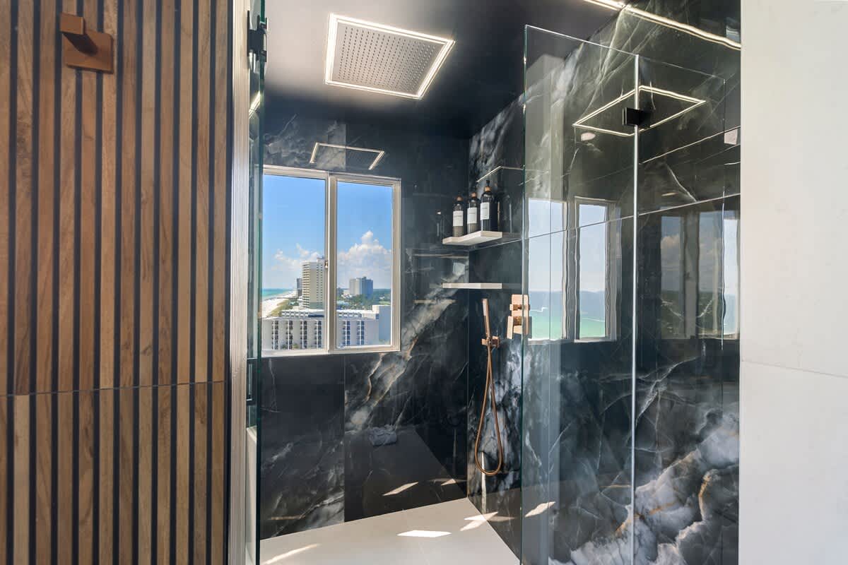 The Summit Penthouse 1501 – Shower area with dark marble walls, modern fixtures, and a view of the coastline through large windows.