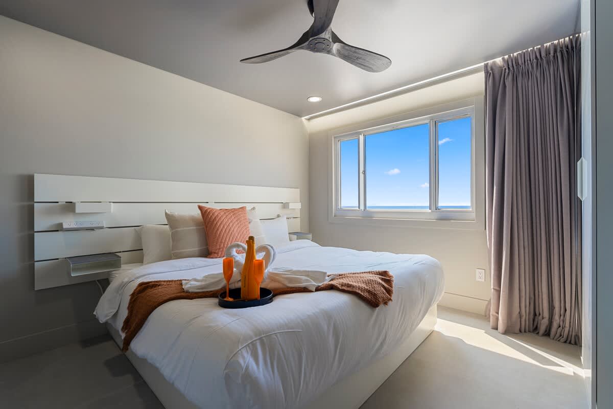 The Summit Penthouse 1501 – Cozy bedroom with a white bed, ocean view, and modern decor, featuring a ceiling fan and large window.