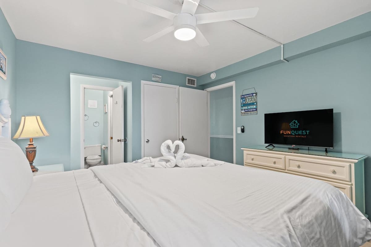 The Summit 315 – Bright bedroom with white bedding, wall-mounted TV, lamp, and attached bathroom door.
