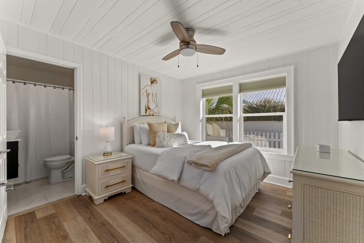 9620 Beach Blvd – Bright bedroom with a queen bed, wooden floors, and en suite bathroom. Windows overlook a garden area.