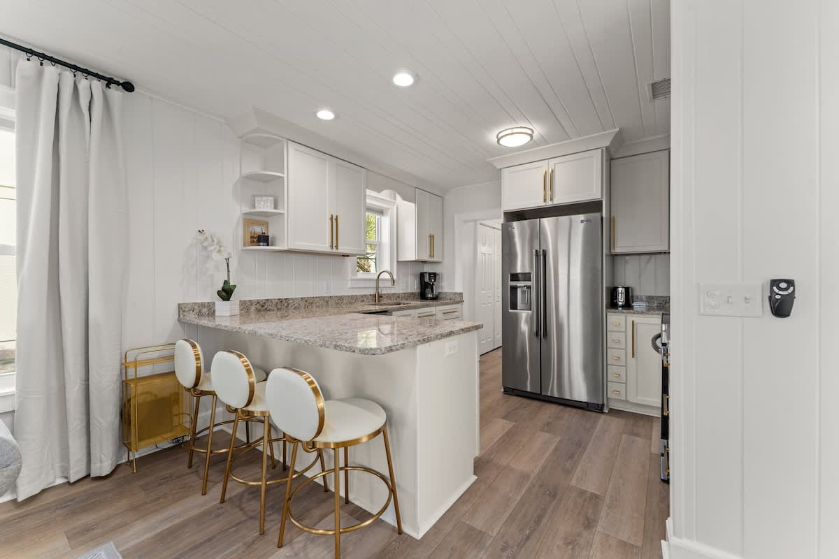 9620 Beach Blvd – Modern kitchen with granite countertop, stainless steel refrigerator, gold-accented bar stools, and white cabinetry.