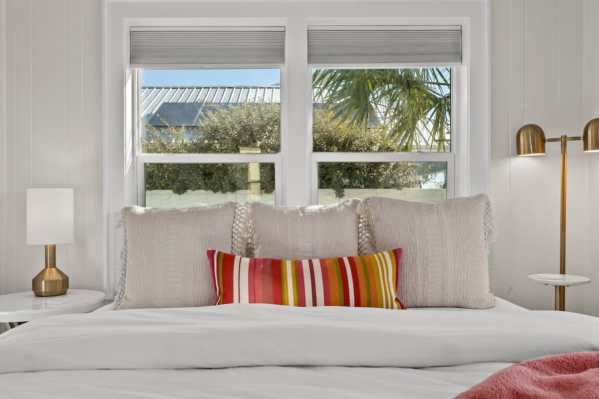 9620 Beach Blvd – Cozy bedroom with decorative pillows, bedside lamp, and windows showing palm trees and a blue sky.