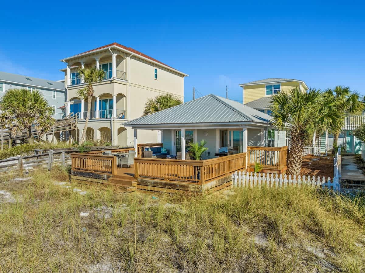 9620 Beach Blvd – Single-story beach house with a deck, surrounded by palm trees and grassy area, blue sky above.