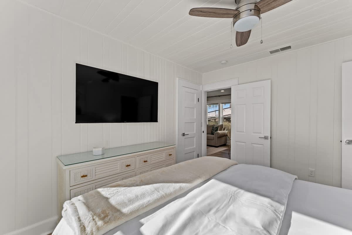9620 Beach Blvd – Bright bedroom with a ceiling fan, TV on the wall, and a view of a living area through open doors.