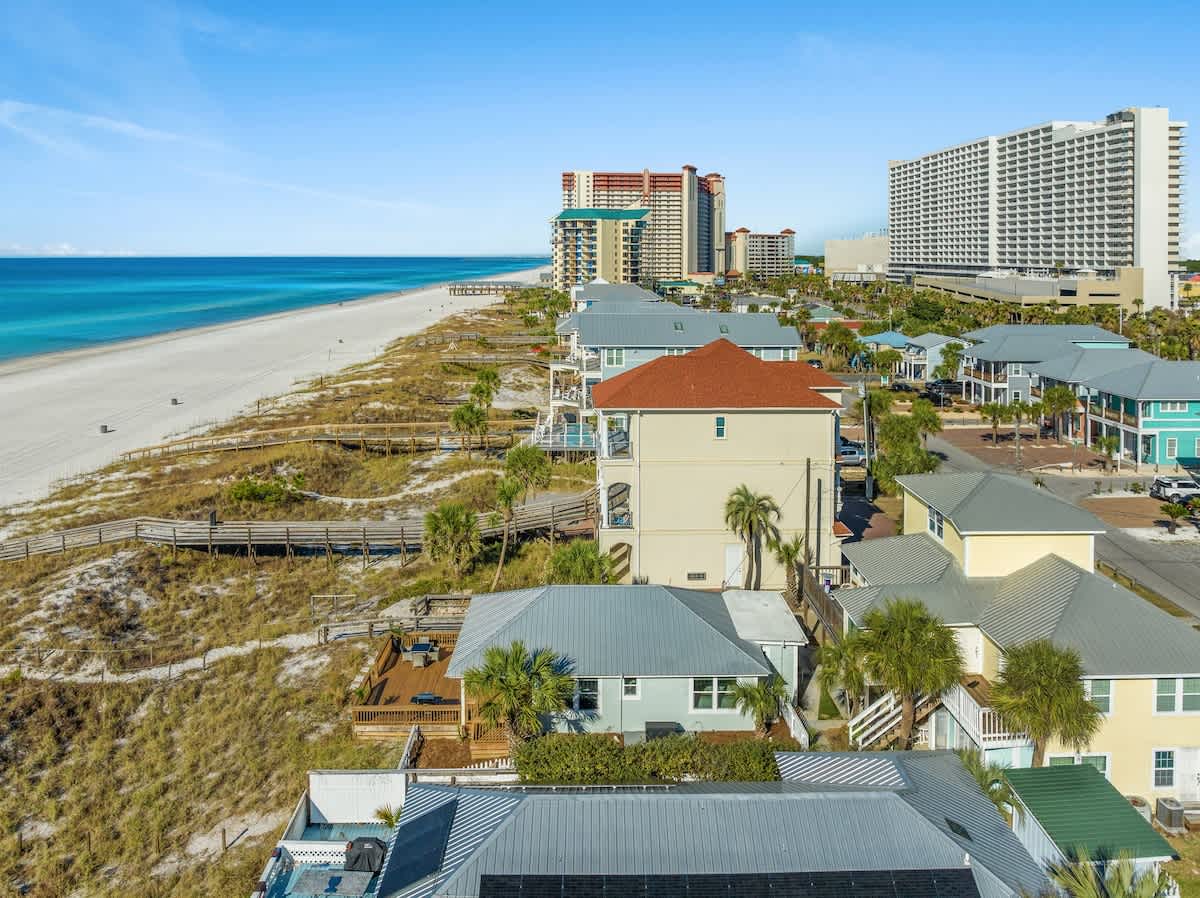 9620 Beach Blvd – Aerial view of beach houses along a sandy shore with turquoise water and nearby high-rise buildings.
