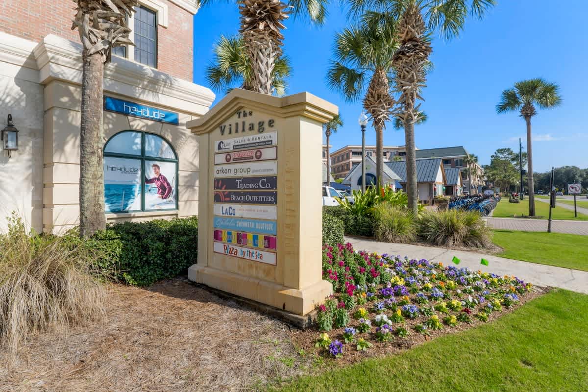Village of SW B257 – Signage for "The Village" with palm trees, colorful flowers, and shops in a sunny outdoor setting.