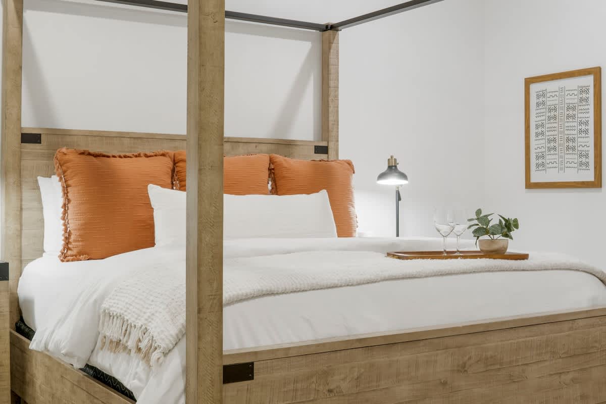 Village of SW B257 – Four-poster bed with orange pillows, white bedding, lamp, decorative tray, and wall art in a bedroom setting.