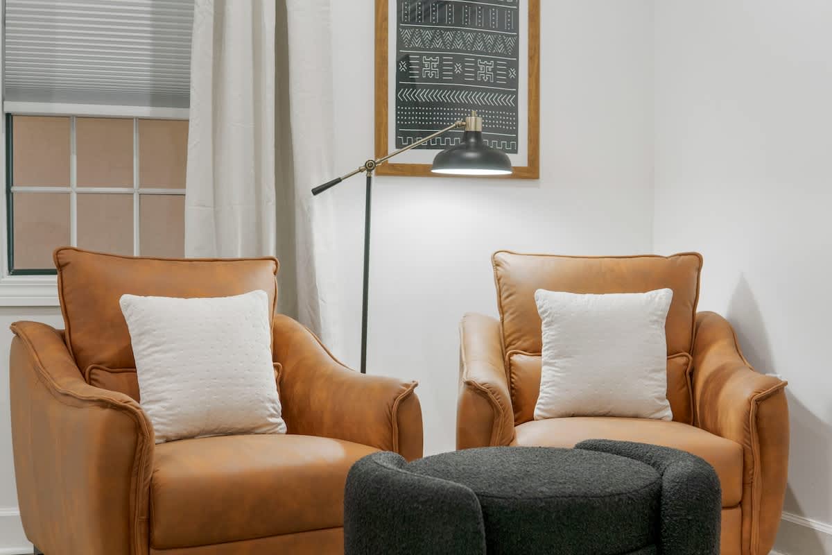 Village of SW B257 – Two tan leather chairs with white pillows and a circular black ottoman, beside a lamp and framed artwork.