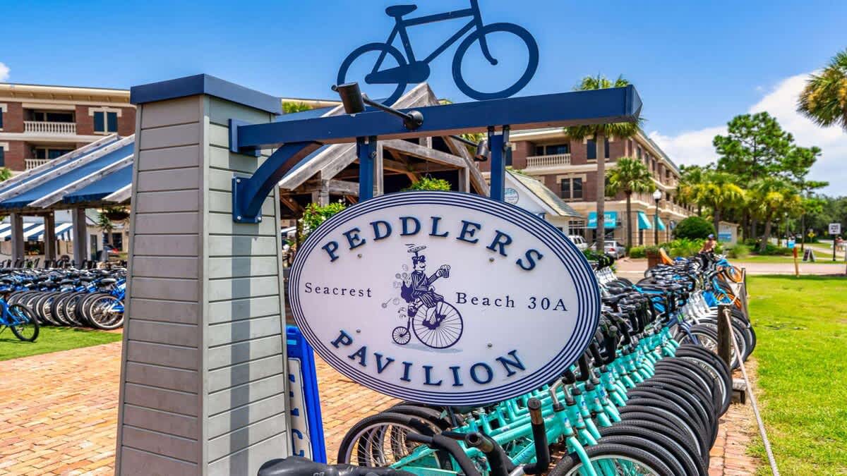 Village of SW B204 – Sign for Peddlers Pavilion with bicycles parked nearby and palm trees in the background.