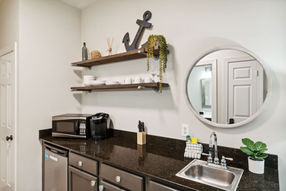 Village of SW B204 – Kitchen area featuring a sink, microwave, coffee maker, shelves with dishware, and a round mirror.