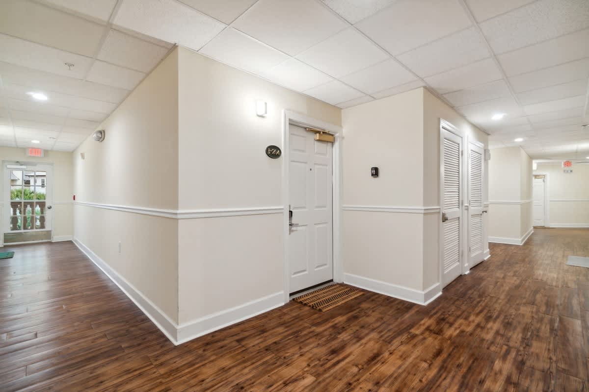 Village of SW B204 – Hallway leading to room entry, featuring light-colored walls and wooden flooring.