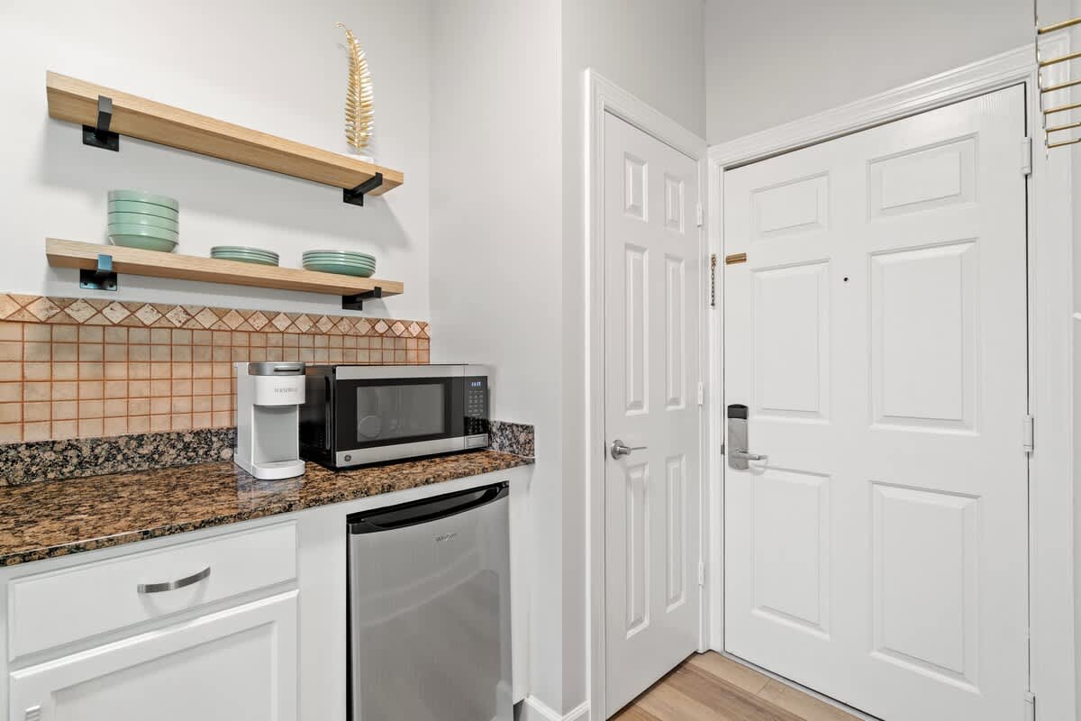 Village of SW B203 – Compact kitchen area with microwave, coffee maker, and open wooden shelves.