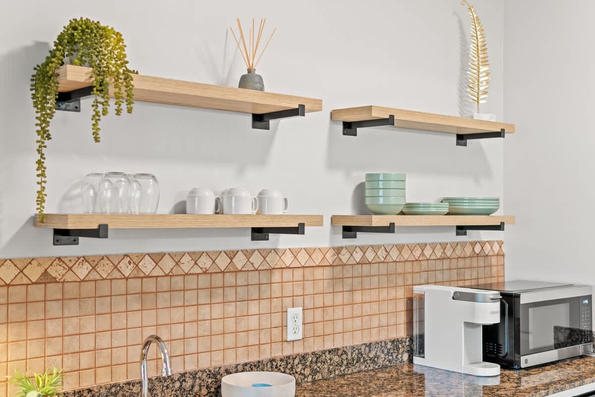 Village of SW B203 – Open kitchen with wooden shelves, glassware, plates, a microwave, and granite countertop.