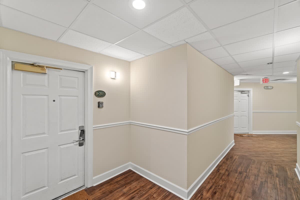 Village of SW B203 – Hallway leading to room B203 with brown laminate flooring and light-colored walls.
