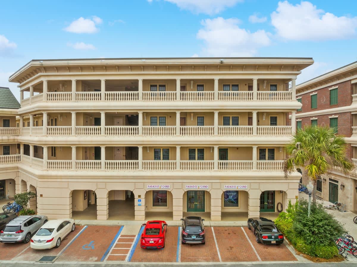 Village of SW B203 – Three-story building with balconies, parking area, and palm trees under a blue sky.