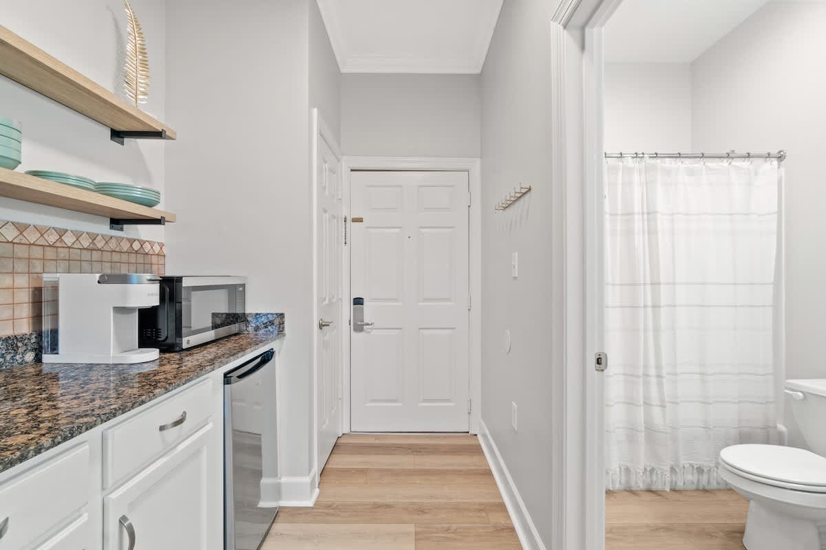 Village of SW B203 – Kitchenette area with microwave and mini fridge, bathroom doorway, light-colored walls, hardwood floor.