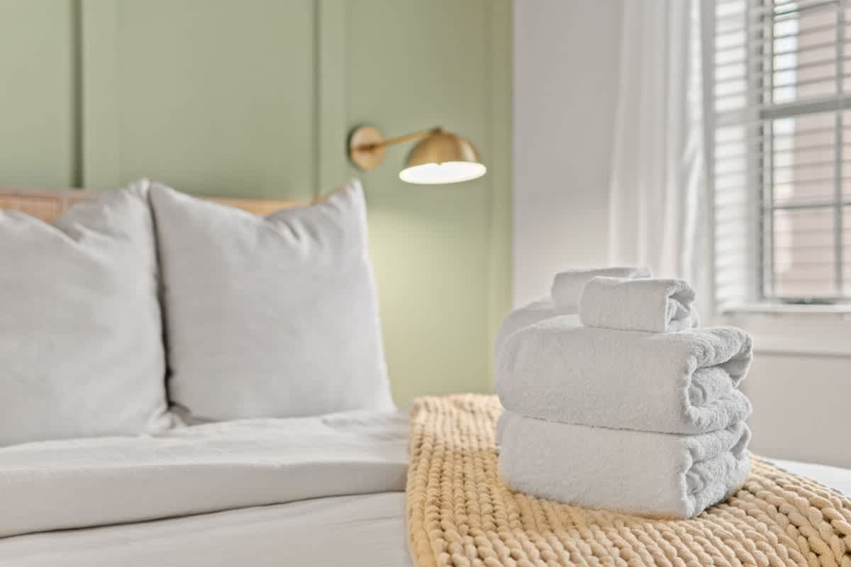Village of SW B203 – Bedroom with green wall, large pillows, stacked towels, and soft yellow blanket.