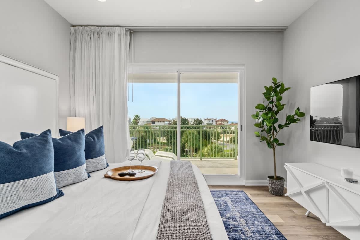 Inn at Seacrest 409 – Bedroom with blue pillows, balcony access, palm trees visible outside, and a television on the wall.