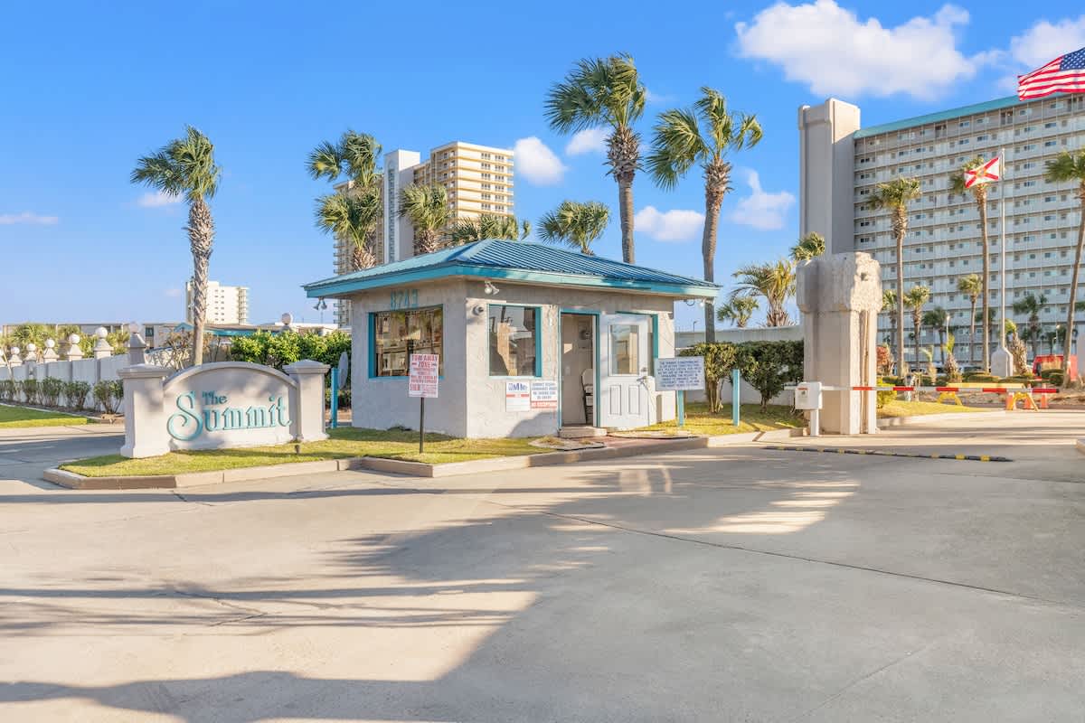 The Summit 1009 – Entrance to The Summit with a guardhouse, palm trees, and nearby buildings under a clear blue sky.