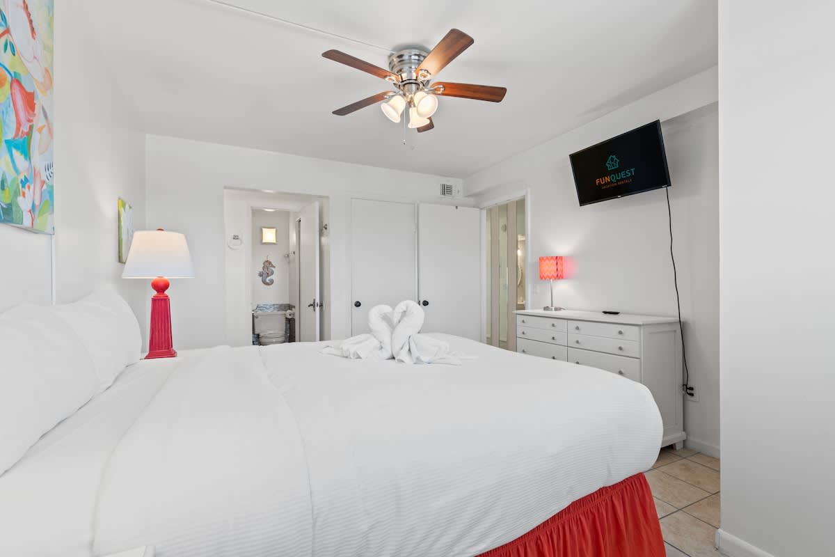 The Summit 1009 – Bright bedroom with queen bed, ceiling fan, TV, and decorative lamp, leading to a bathroom.