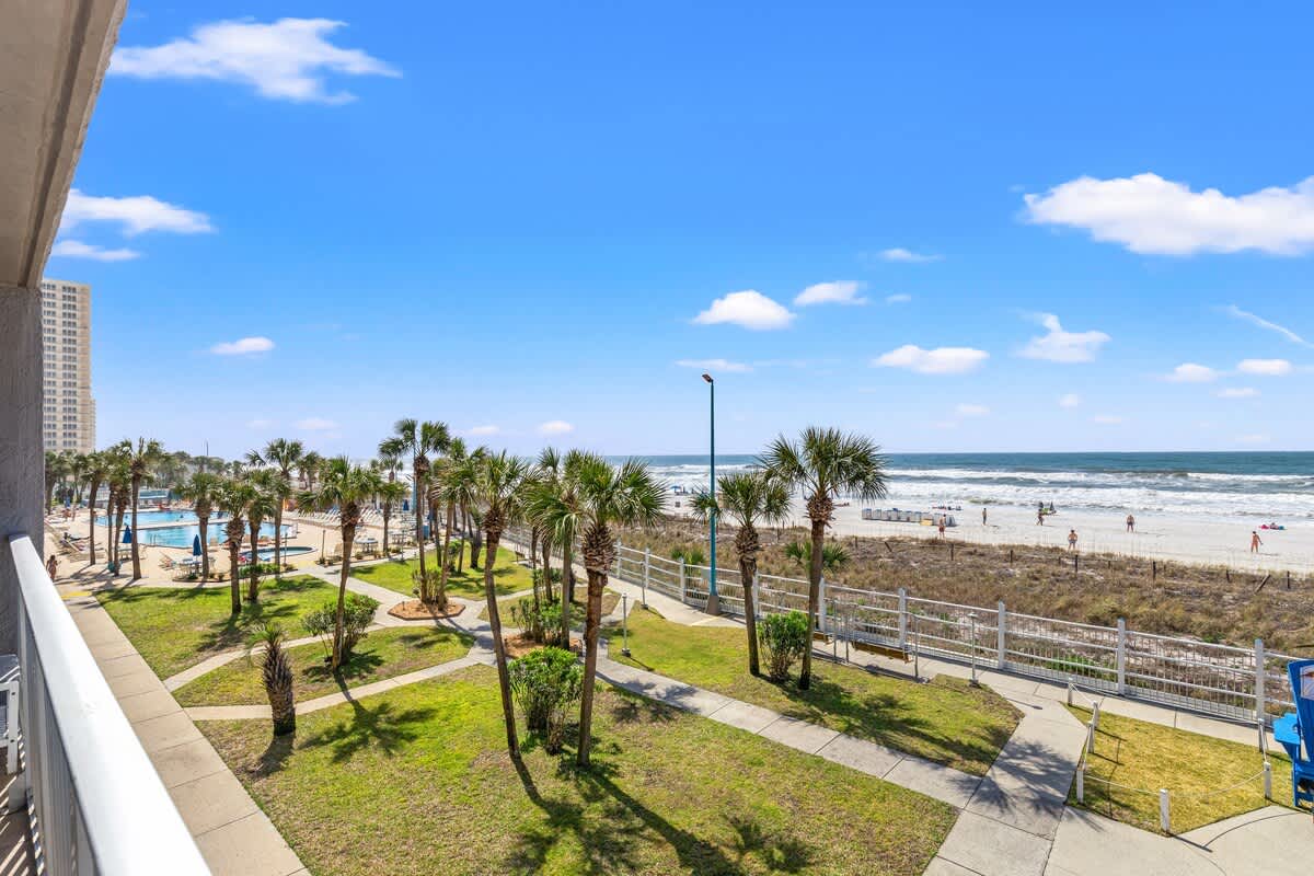 The Summit 301 – View from balcony showing palm trees, lawn area, pool, and beach with ocean waves in the background.