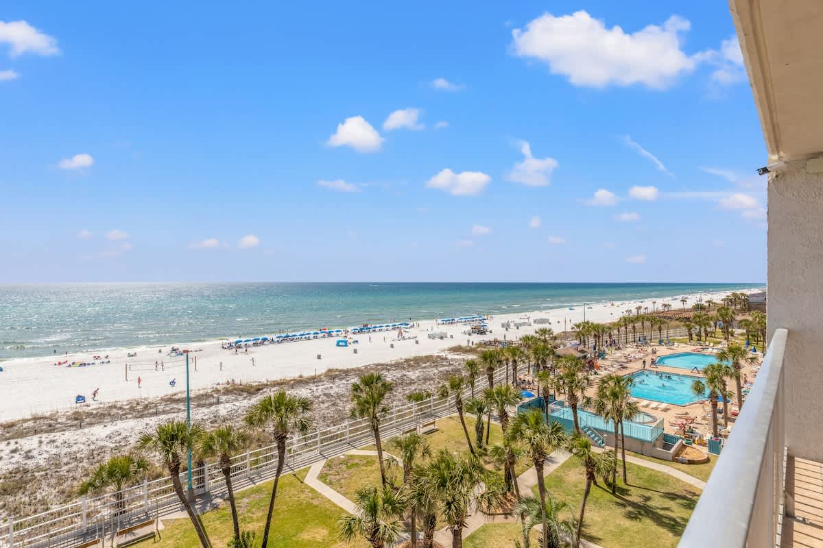 The Summit 632 – View from balcony showing pools, palm trees, and beach with umbrellas under a clear blue sky.