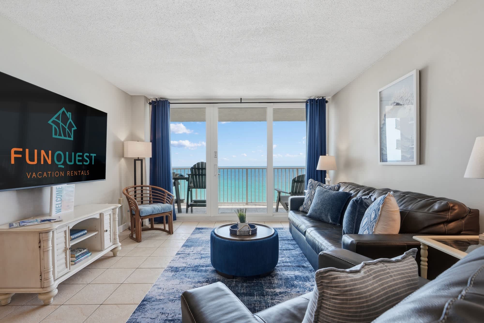 Dunes E1702 – Living room with ocean view, brown leather sofa, blue rug, and balcony access.