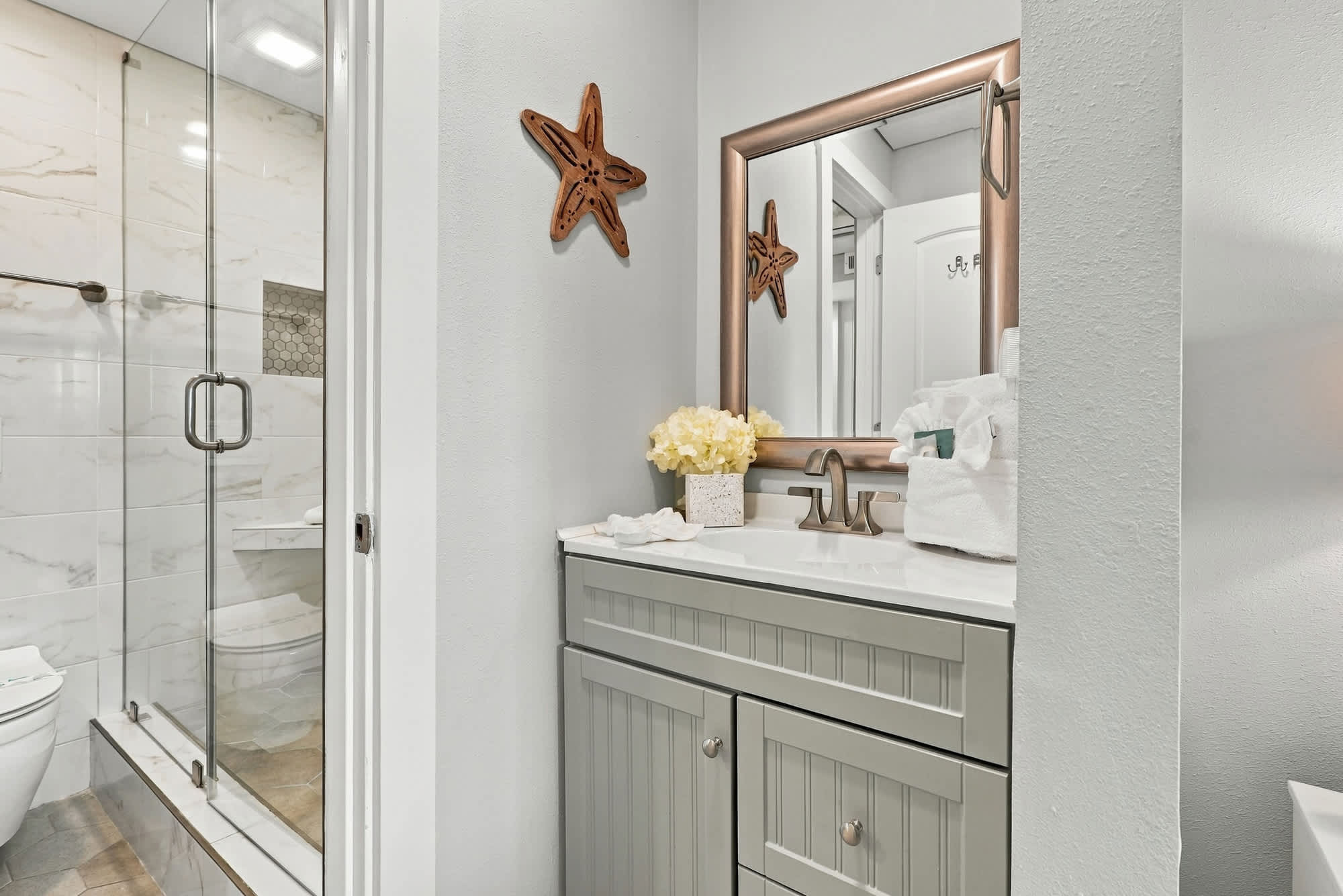 The Summit 1122  – Bathroom with grey vanity, mirror, starfish decor, and glass shower. White towels and flowers on the counter.