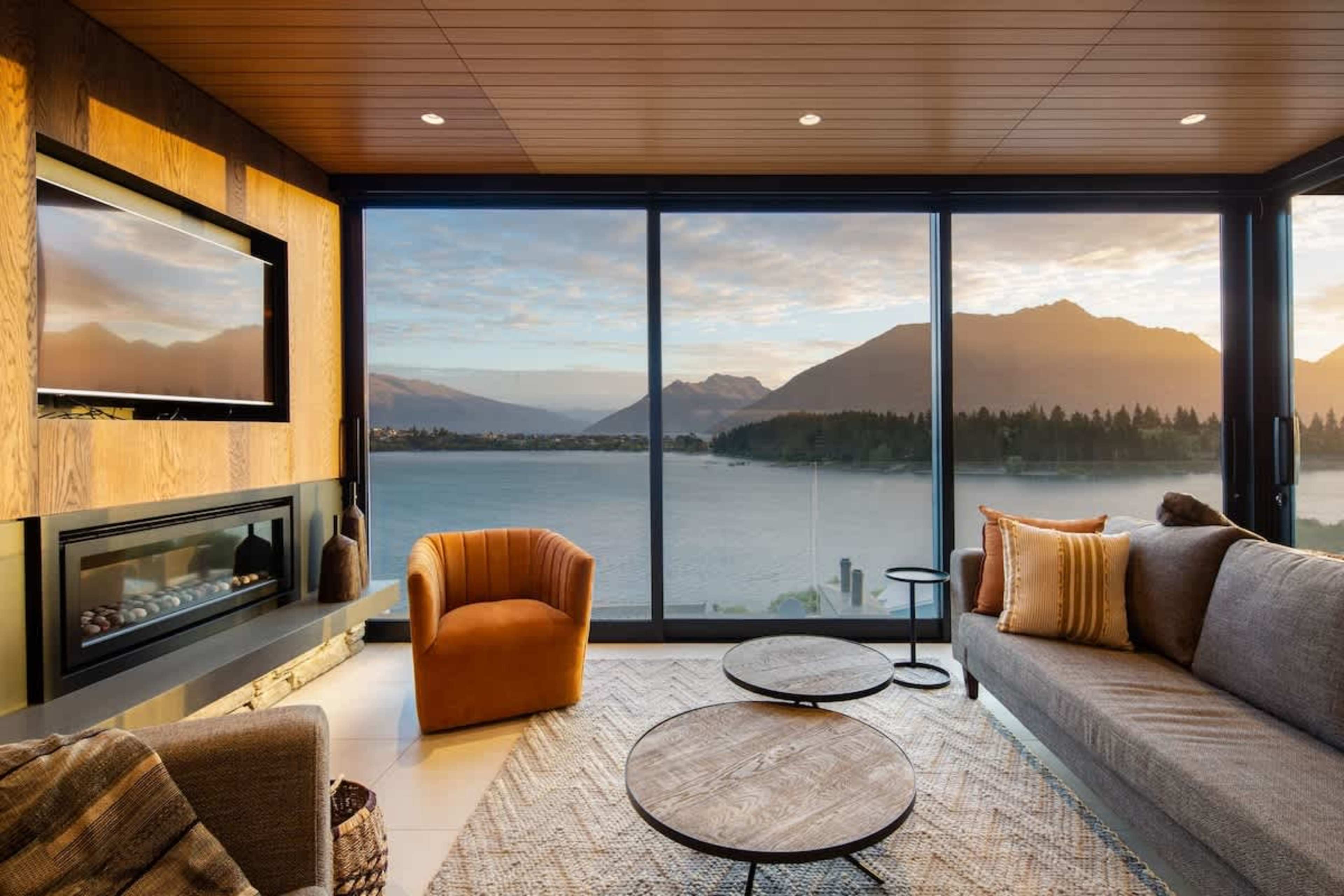 Experience elegance in this Queenstown villa's living room, featuring contemporary furnishings, a statement fireplace, and a private terrace. Enjoy panoramic views of Lake Wakatipu and the stunning surrounding mountains.