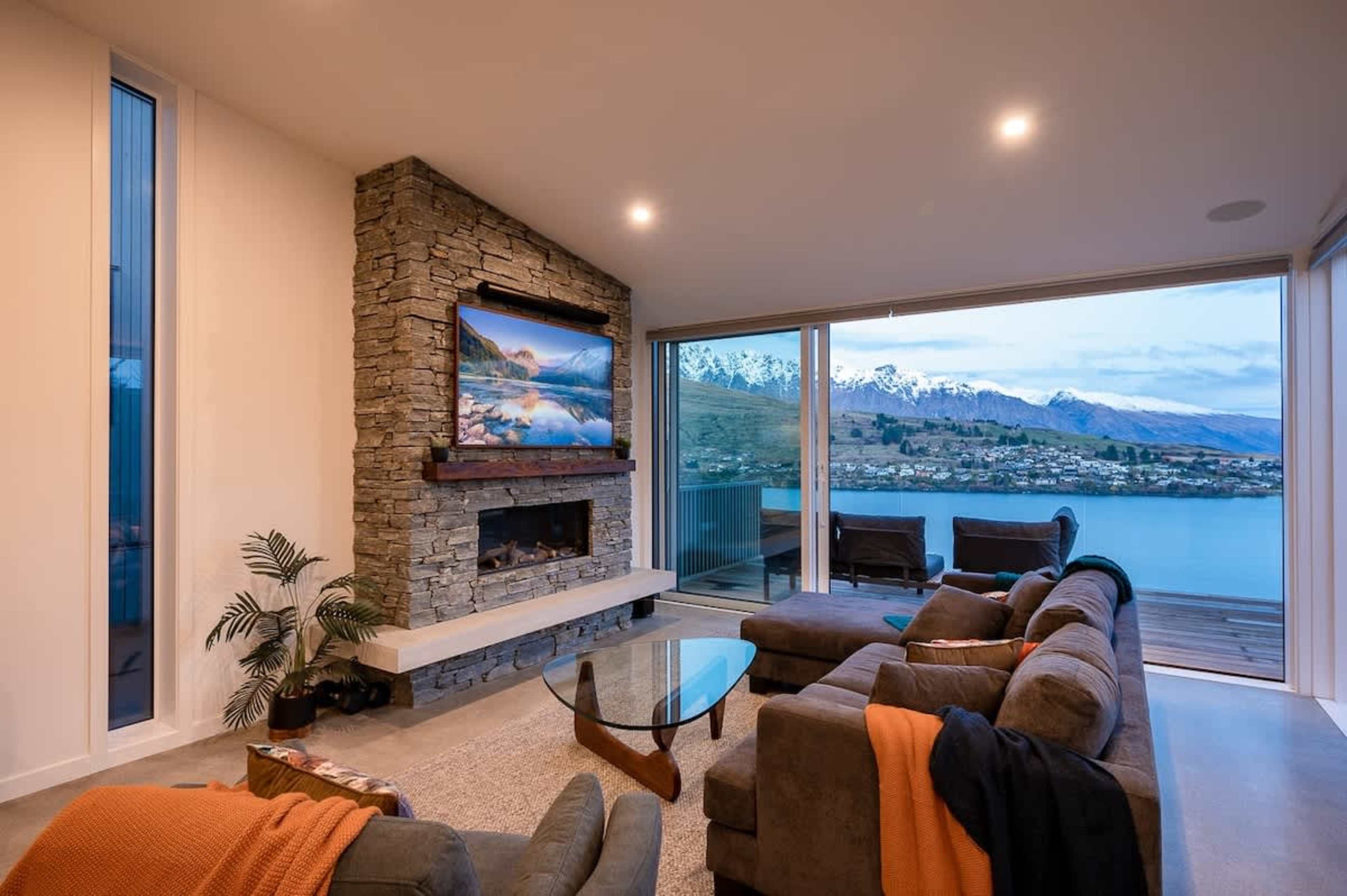 The spacious living room of this 4-bedroom villa features an inviting gas fireplace, comfortable seating, and large windows offering panoramic views of Lake Wakatipu and the surrounding mountains.